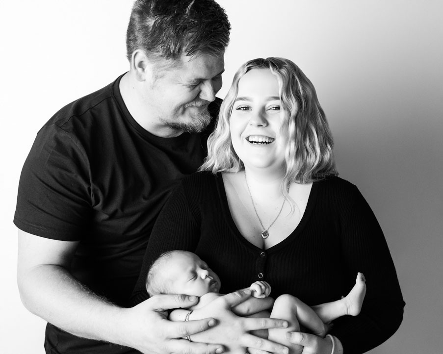 Black and white photo of mum and dad holding baby smiling portrait at Kristy’s Studio, Blue Mountains — relaxed, joyful moment